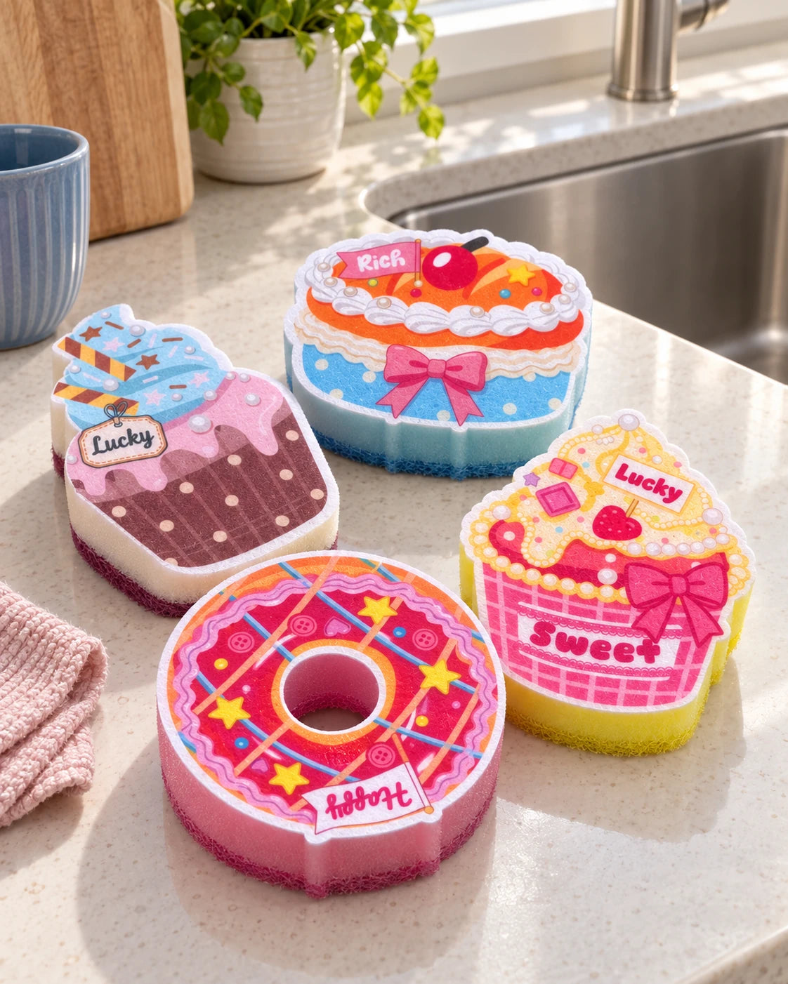 Four dessert-shaped kitchen sponges arranged beside a stainless-steel sink: a pink donut, layer cake and two cupcakes.
