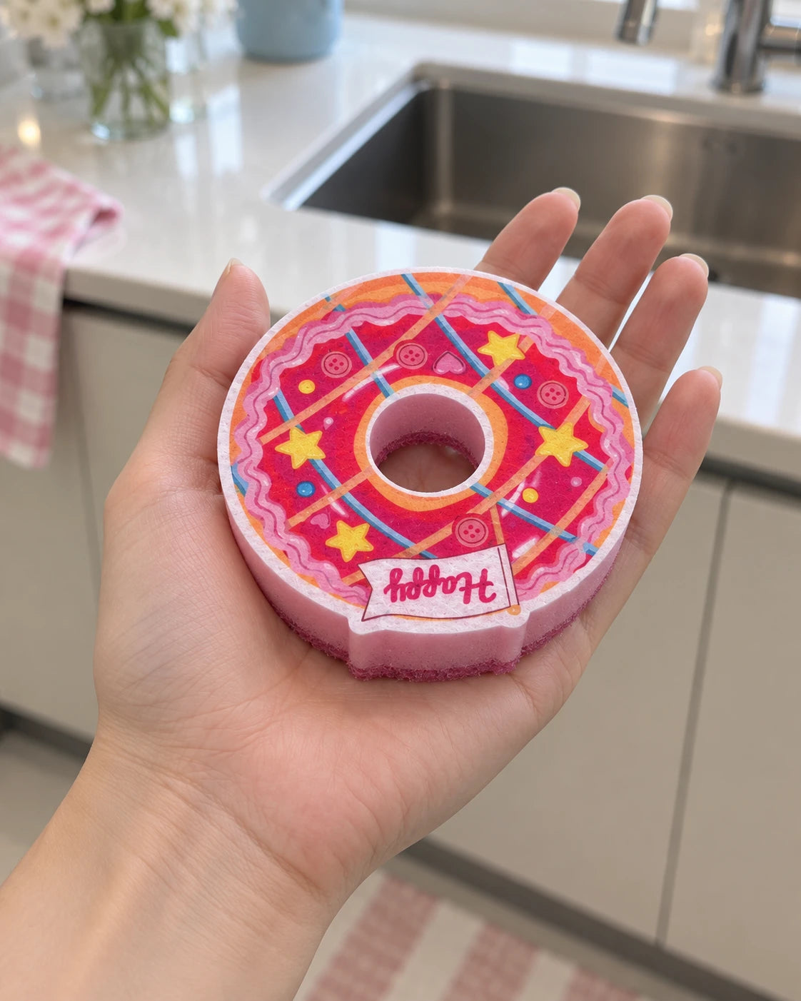 The pink ring donut kitchen sponge rests in an adult palm, showing its open center, printed design and relative scale.